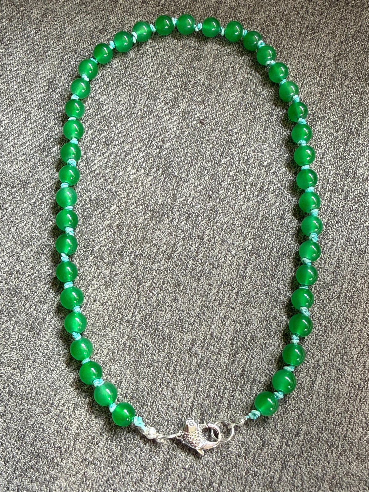 A beaded necklace made of glossy green round beads with small light blue knots and a silver fish-shaped clasp.